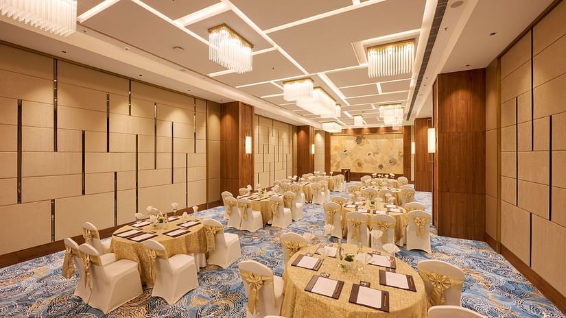 A formal event space is prepared with round tables covered in gold cloths, each with place settings and surrounded by chairs adorned with gold sashes, under a modern ceiling light design. - Iris Sarovar Portico, Sector 85, Gurugram