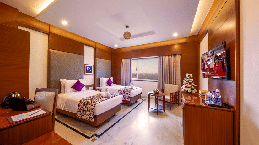 A well-lit Executive Room at JP Hotel Chennai featuring twin beds facing a wall-mounted TV, wooden furnishings and a window overlooking the view outside.