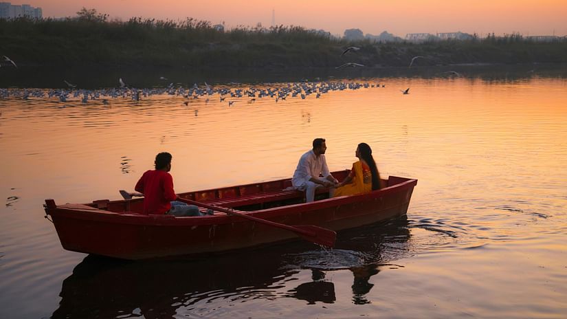 Two people rowing a boat on a calm lake at sunset with birds flying in the distance.