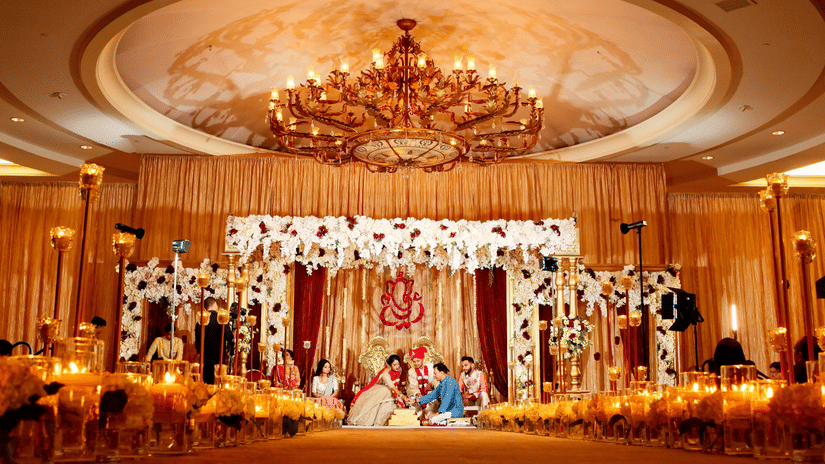 An indoor Indian wedding ceremony on a lavishly decorated stage, with the couple seated together and surrounded by their family.