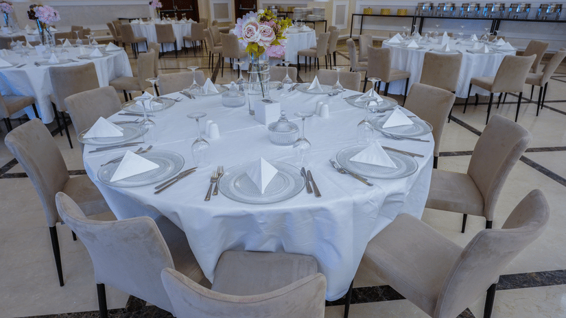 A banquet hall with round tables set for an event. Each table is covered with a white tablecloth and arranged with plates, napkins, and cutlery. Neutral-coloured chairs surround the tables, and a floral centrepiece adorns each at La Maison Hotel, Doha.