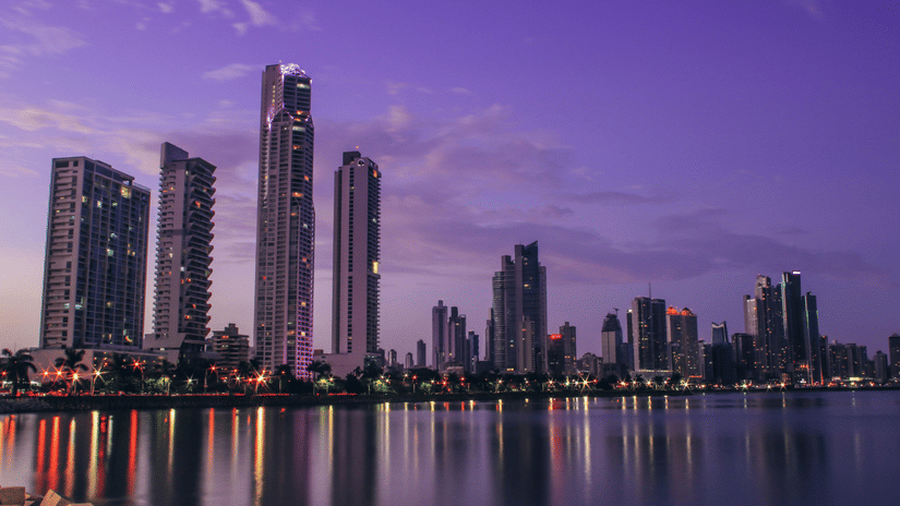 A purple-hued sunset over a city skyline with tall buildings reflected in the calm water below.