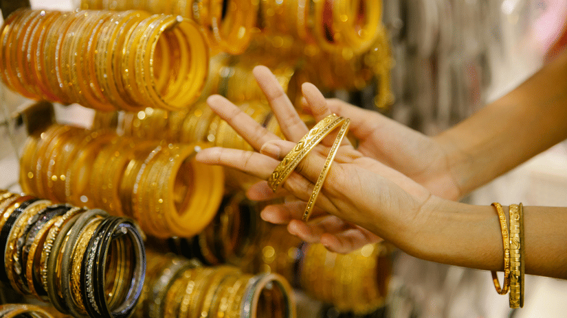 A person's hands are shown trying on multiple gold-coloured bangles from a large collection hanging on a stand.