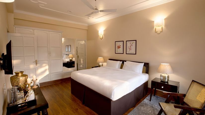 An image of super deluxe room featuring a king size bed, bedside table on either side, a flat TV screen, a table where antique items are placed, and  a chair in one corner of the room- Lamrin Boutique Cottages Rishikesh