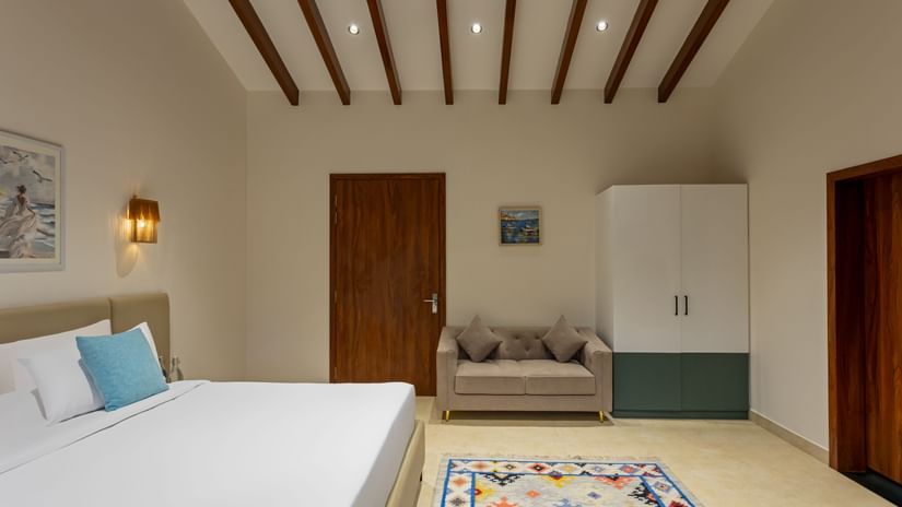 Spacious bedroom with wooden ceiling beams, seating area, wardrobe, and minimalist decor – Lamrin Morjim Goa