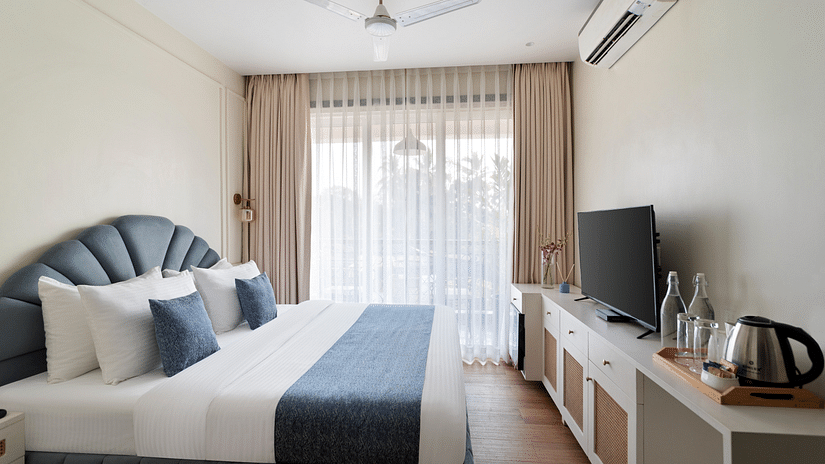 A well-lit daytime image of a room featuring a stylish grey bed, a storage cabinet, a TV, a tea and coffee maker, a ceiling fan, an air conditioner, and a large window.