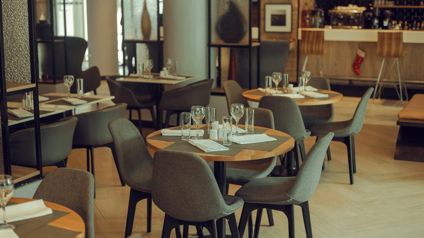 An indoor dining space with round wooden tables and grey upholstered chairs featuring a bar area and large windows with natural light at the background.