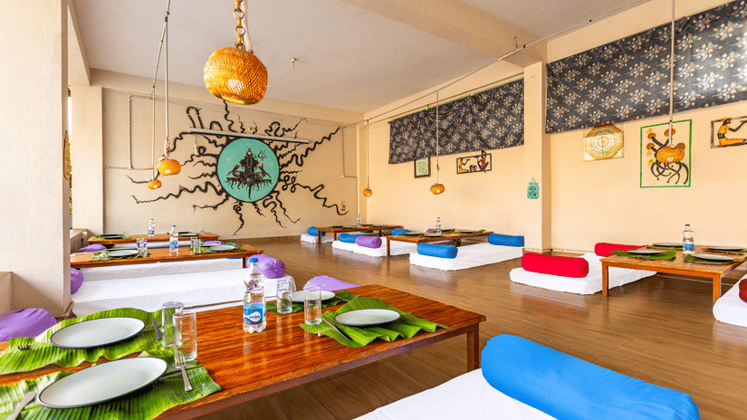 Low-angle view of a restaurant table set with banana leaves and steel plates, featuring colorful bolster cushions - Nature Trails Ashoka Resort Hampi
