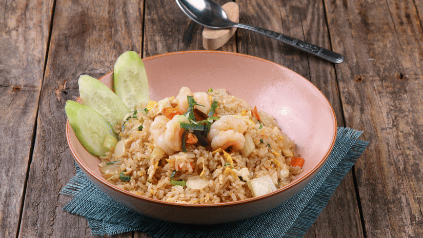 Top-down view of a Thai-style rice dish served on a peach-coloured plate with cucumbers on a wooden table.