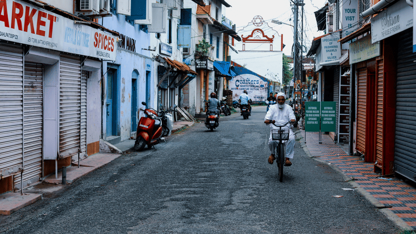 A quiet heritage street in Fort Kochi in monsoon with shuttered shops, whitewashed buildings and a cyclist riding through the narrow lane beneath cloudy skies