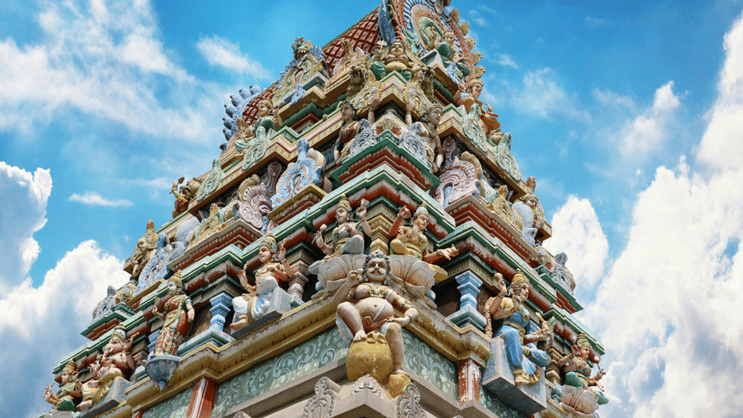 A richly decorated, colourful Hindu temple gopuram (tower) against a bright blue sky with clouds.