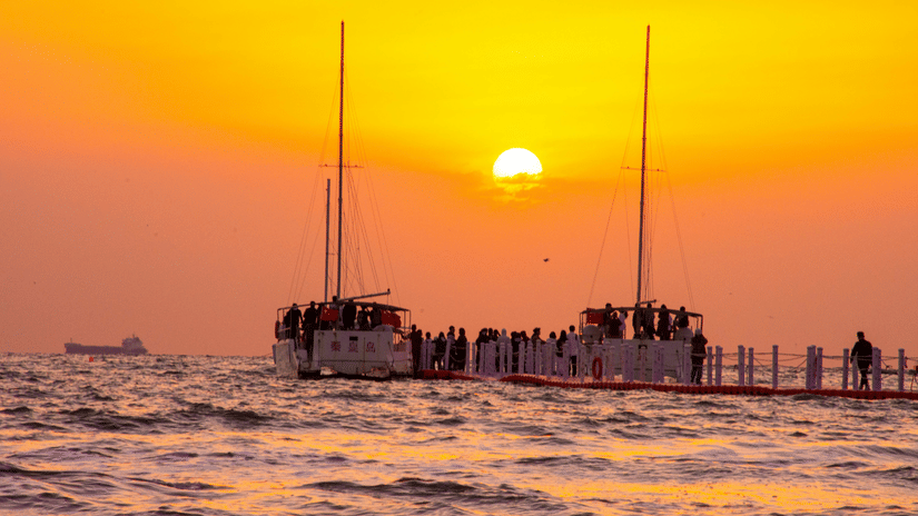 A vibrant orange sunset over the sea, with 2 boats docked at a pier packed with people viewing the scene.