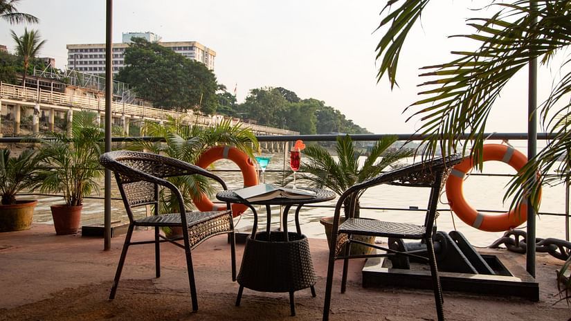 A waterfront outdoor seating area with wicker chairs, a table, and lifebuoys, overlooking a river with a cityscape in the distance. - polo floatel kolkata