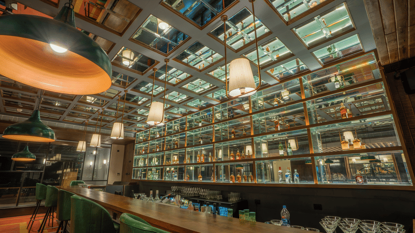 Bar at Polo Hotel, Agartala with backlit mirrored bottle display, green pendant lights, copper accents and green leather bar stools at long wooden counter.