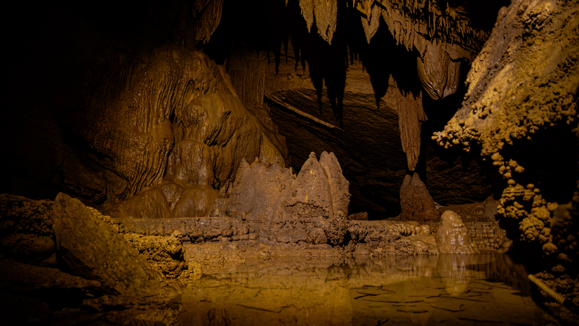 Warmly lit cave chamber with stalactite and stalagmite formations rising above a calm reflective pool inside a dark underground limestone cavern.
