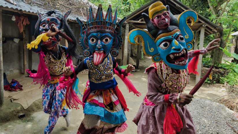 Three Khasi performers in colourful tribal masks and ribboned costumes dancing in a traditional cultural performance in a Meghalaya village.