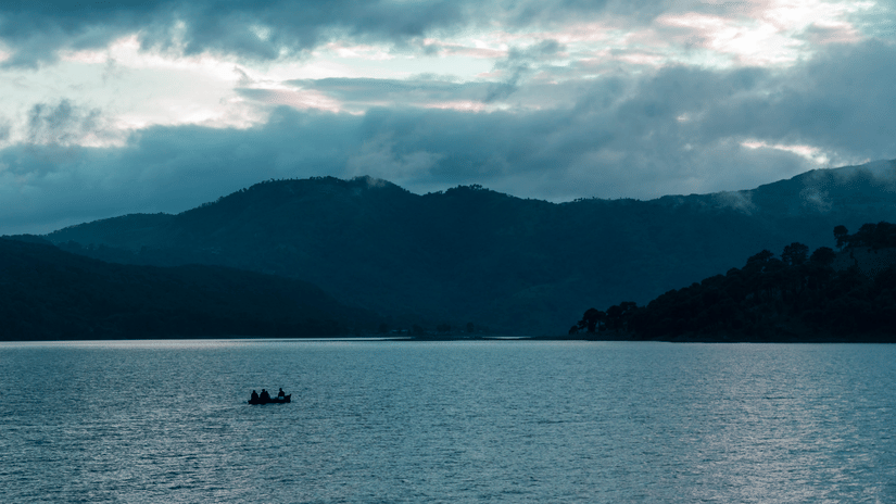 Two silhouetted people in a small boat on a calm lake with misty forested hills and dramatic cloudy sky over Meghalaya at dusk.