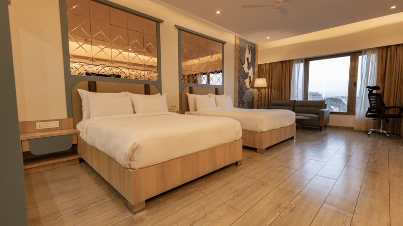 A well-lit hotel room featuring twins beds, a sofa near a window, wooden flooring, and a patterned headboard or wall panel | Pride Elite Chhatrapati Sambhajinagar
