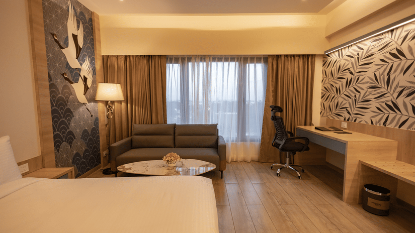 A well-lit hotel room with a large bed in the foreground, a sofa, a work desk, and a decorative wall panel with a leaf pattern | Pride Elite Chhatrapati Sambhajinagar