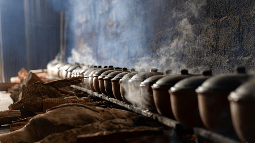 A row of clay pots sits over a fire in a dark indoor space. Steam rises from the pots into the air.