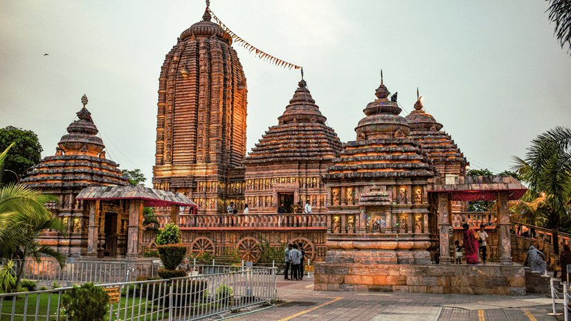 A stone temple with multiple tiered spires and carved walls stands under a bright sky. A paved walkway leads toward the entrance where people are gathered.