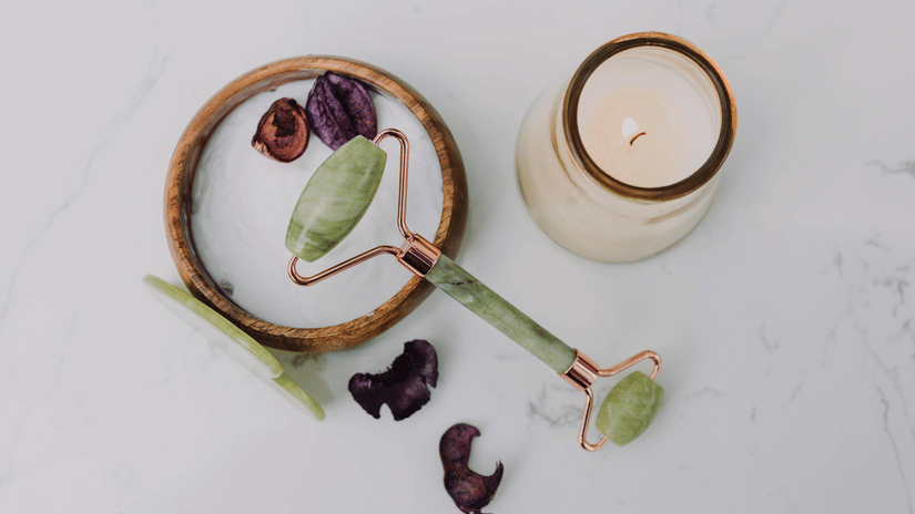 A jade roller lies on a white surface next to a lit candle and dried petals.