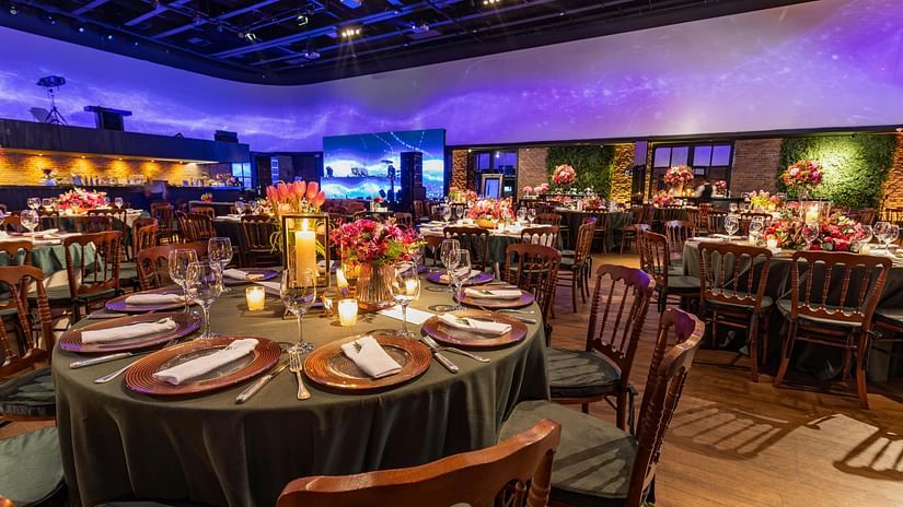 A wide shot of a large indoor venue set up for a formal event or gala dinner, with numerous round tables, chairs, and ambient lighting.