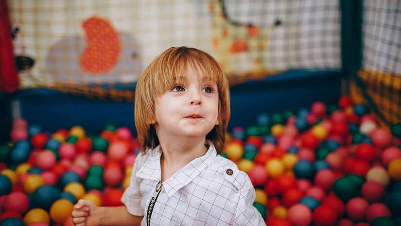 A young child with blond hair is standing in a colourful ball pit, looking with a slight smile.