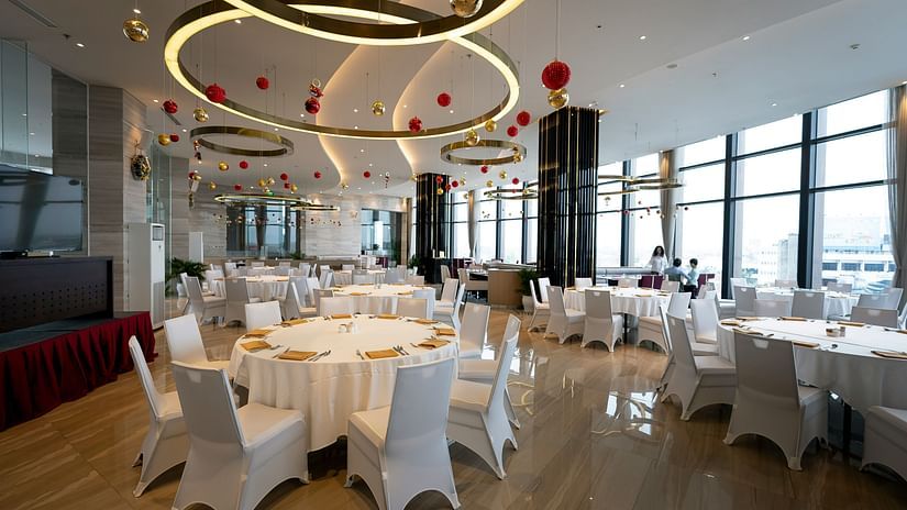 A bright, modern event hall or restaurant space set up with round tables, white chairs, and elegant ceiling lights, with large windows overlooking a city.