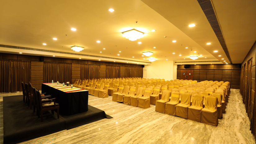 A view of the theatre style seating arrangement at one of the best wedding halls inside Raj Park Hotel, Chennai, featuring chandeliers, a stage and a podium.