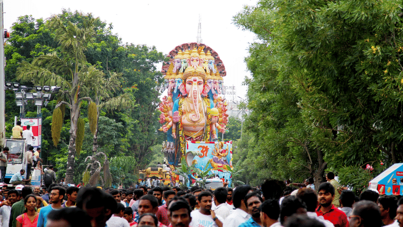 Devotees gathering around a towering Ganesha idol during a procession for Vinayaka Chaturthi festival, with colourful decorations and greenery lining the route.