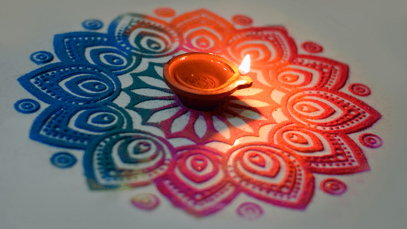 a rangoli made by hand with different colours and a diya lit up in the centre of the rangoli