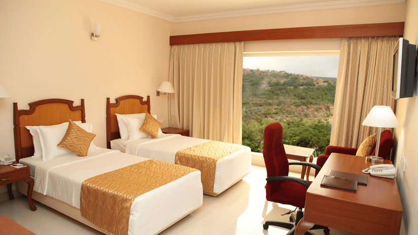 One of Raj Park Hotel's superior rooms in Tirupati featuring twin beds with gold runners, a wooden desk with a red chair, and a large window overlooking a forested hillside.