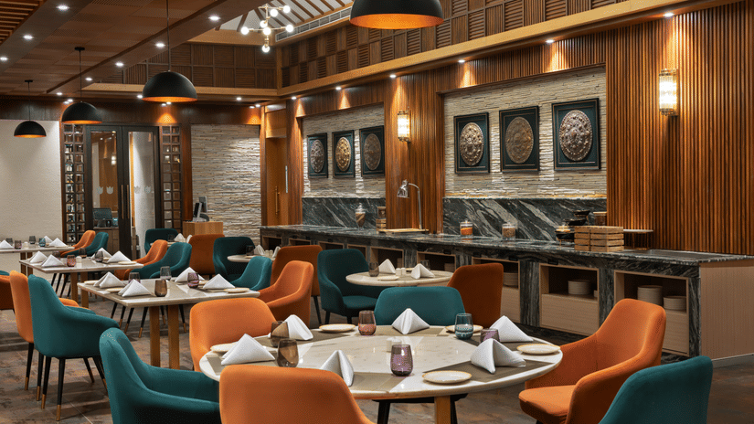 Guras restaurant interior at Royal Tulip Shivapuri National Park - Kathmandu featuring round tables, orange and teal chairs, and wood panelling.