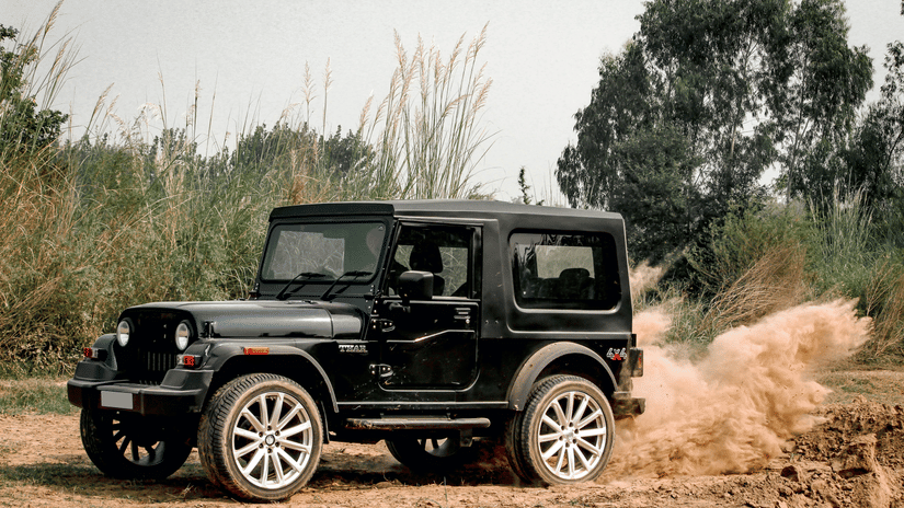 A black Mahindra Thar SUV drives on a dirt road, kicking up a cloud of dust behind it. 