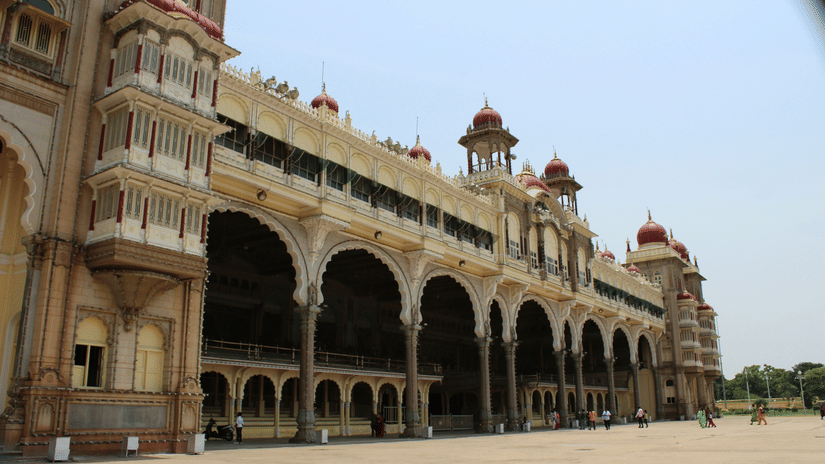 A view of a magnificent palace with arches on the side of the entrance and large open space in front and people walking on it.