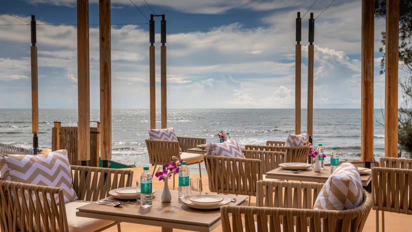 A well arranged tables and chairs at In-House Restaurant with sea waves crashing on the beach in the background