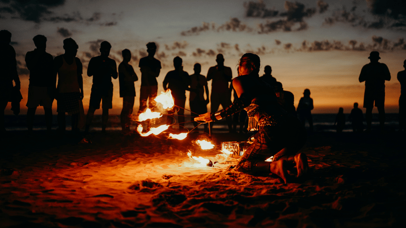 A group of people gathered around a bonfire on a beach at sunset.