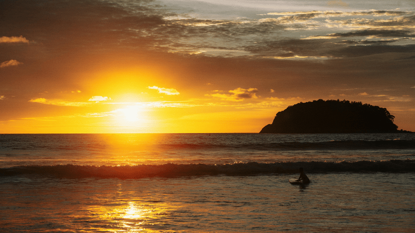 A vibrant sunset illuminates the sky and ocean, with a dark island silhouette visible in the distance.