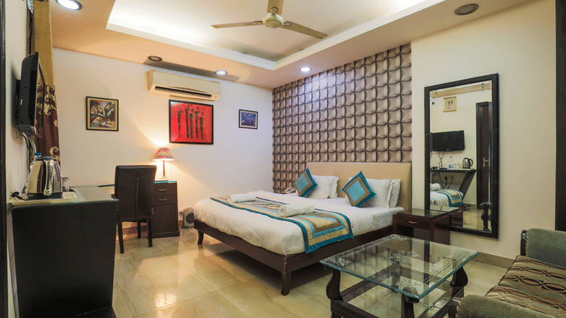 A bedroom featuring a bed, a desk with a chair, a glass table, a sofa, and a mirror that reflects a television.