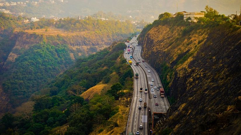 A winding highway, filled with cars, snakes through a lush, green valley framed by steep, rocky hills.