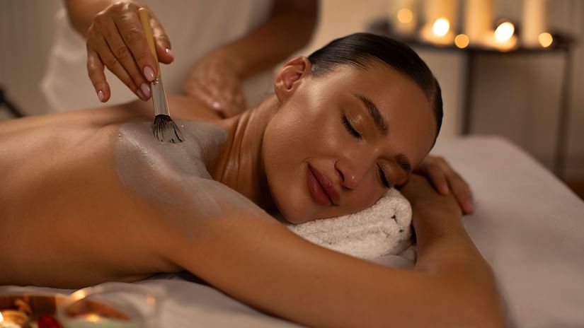 A person receives a body treatment, with a brush applying a mask to their back in a spa setting
