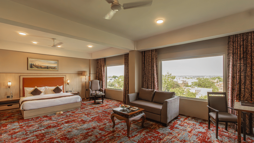 The Junior Suite Room at The Lalita Grand with a double bed, ceiling lights, a 2-seater sofa, ceiling lights, and a window overlooking the greenery.