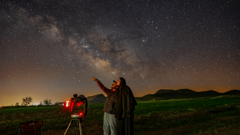A man showing a star to a woman while standing next to a telescope, which is pointed at the milky way behind them.