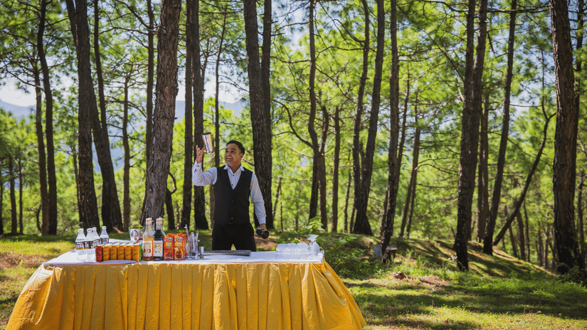 A server preparing drinks at a beautifully draped outdoor buffet table set amidst a lush pine forest - The Manor Sports & Wellness Hotel