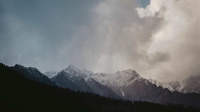 A dramatic view of snow-capped mountains under a stormy, cloudy sky with rays of sun breaking through.