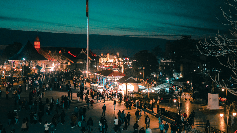 An aerial view of The Ridge in Shimla with many people walking around the shops, a pole with the Indian flag on it and mountains in the background as seen during twilight hour. When deciding the best time to go to Shimla, think about what you are planning 