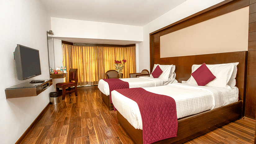 Executive Room at The Monarch Hotel, Brigade Road, Bangalore, with a wooden bed, maroon cushions, a wall mounted television and polished wooden flooring.