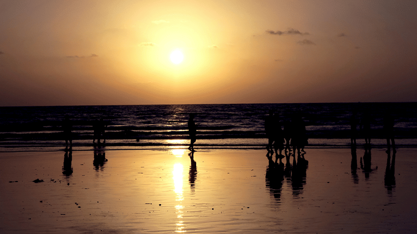 A view from the shoreline of the Aksa Beach sunset, with numerous people standing on the shore and waves overlapping it