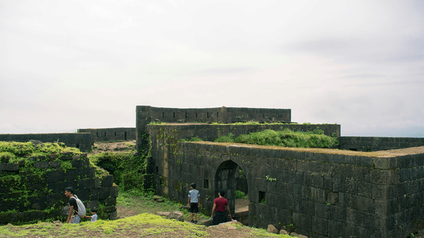 A far-out view of a fort, symbolising Madh Island Fort, that is being consumed by greenery with people also in view.